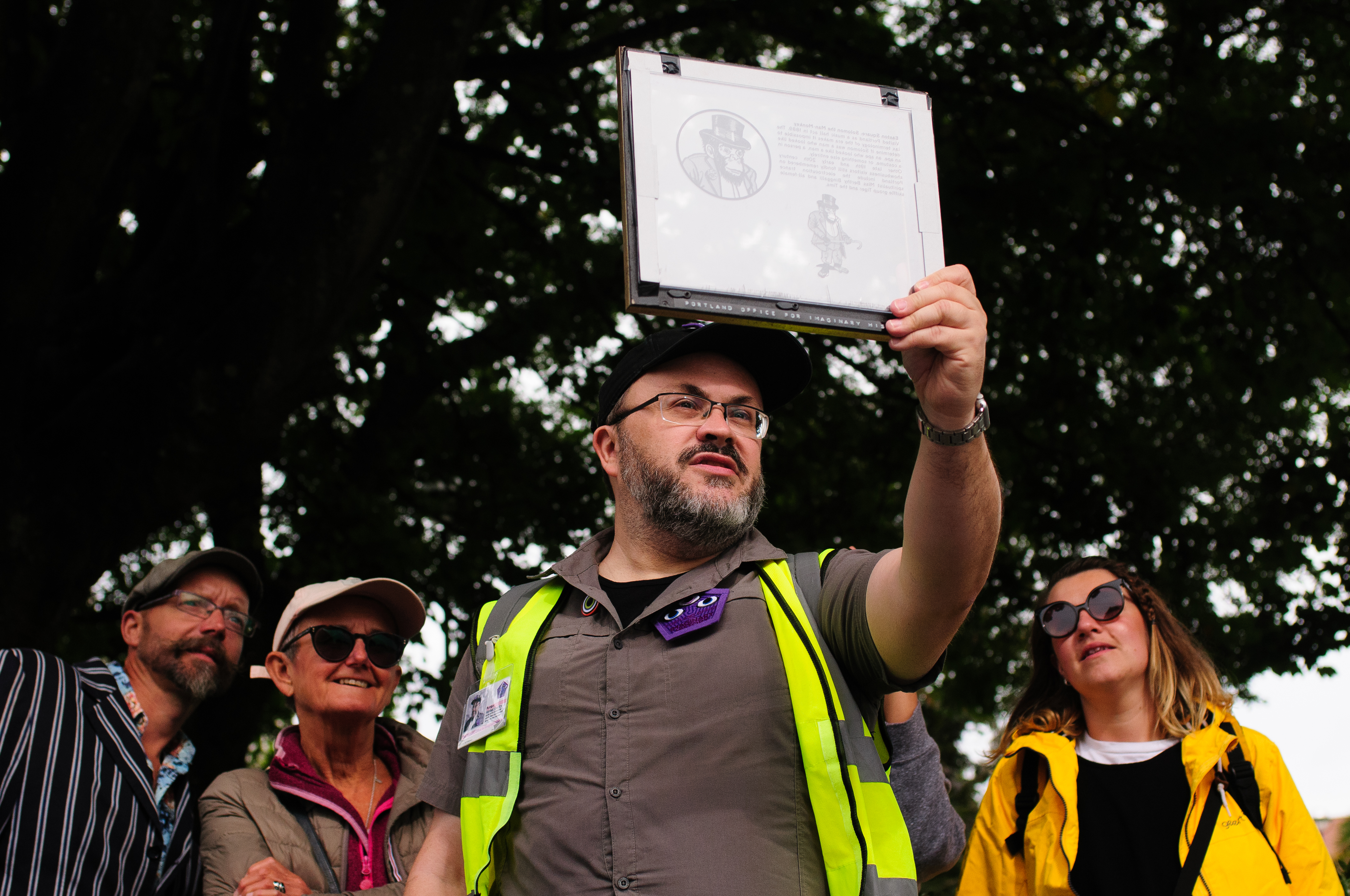 Alistair Gentry in uniform as an imaginary history ranger (khaki shirt, cap and yellow hi vis jacket), leading a tour and showing an image through a transparency viewer.