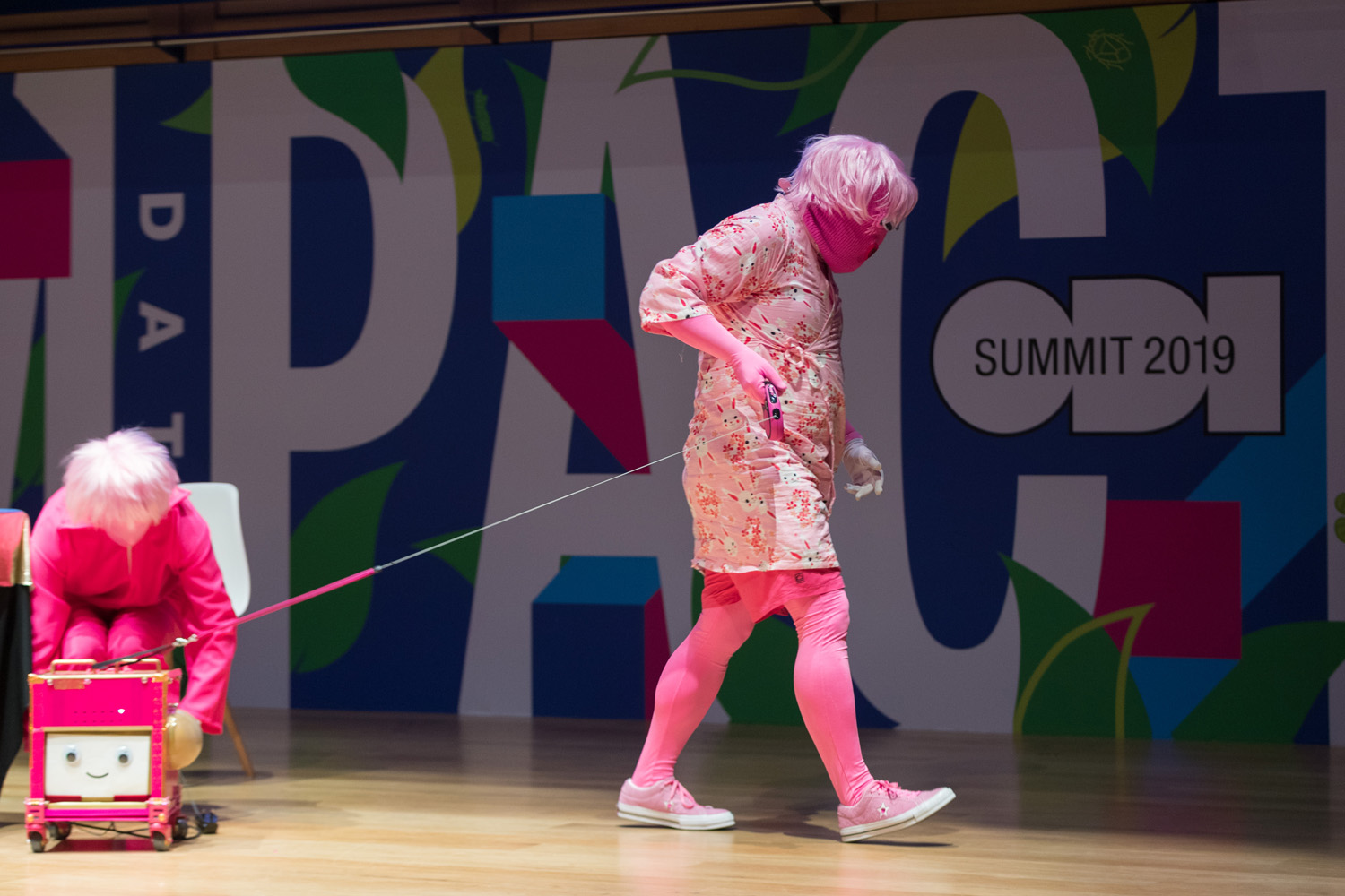 On a stage, a person dressed in pink, including a pink mask and pink wig, walking a pink robot with smiling face on a pink dog lead.