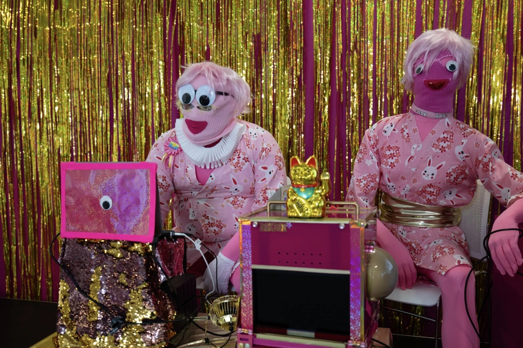 Two people dressed in pink, with pink balaclavas and pink wigs, operating hot pink and gold electronics in front of a gold curtain.